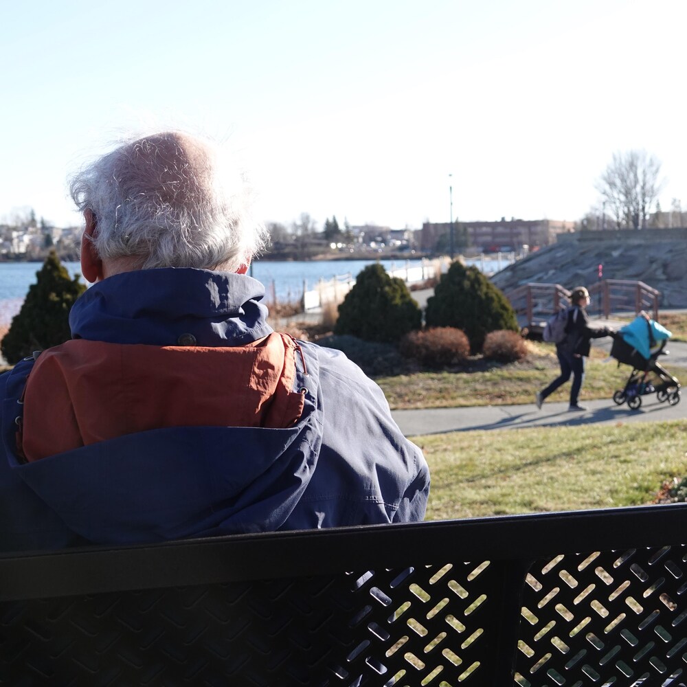 Pierre Vincelette, de dos, est assis sur un banc et il regarde vers le lac.