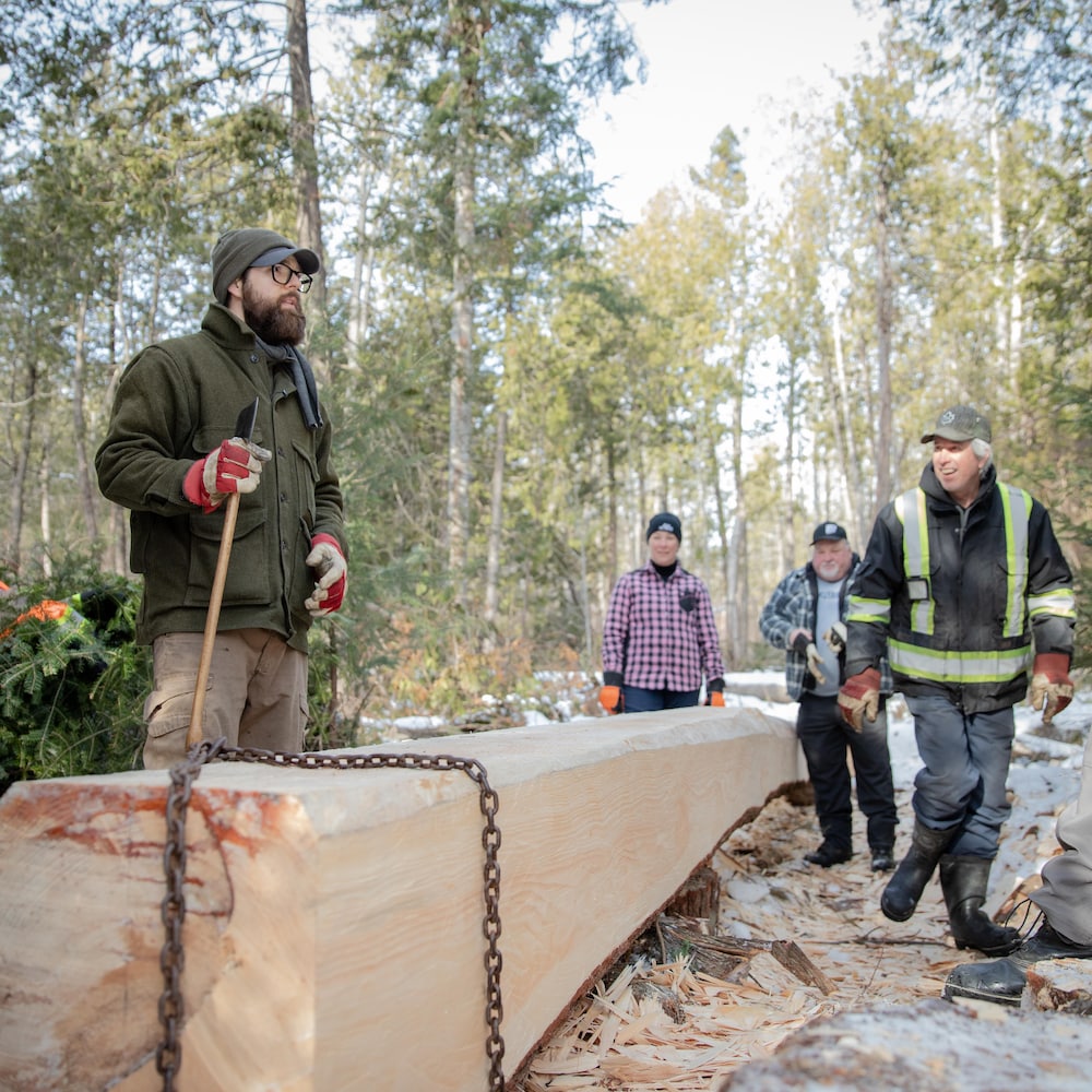 Un groupe de personnes entoure une longue poutre de bois équarrie et ceinturée d'une chaîne dans une forêt enneigée.