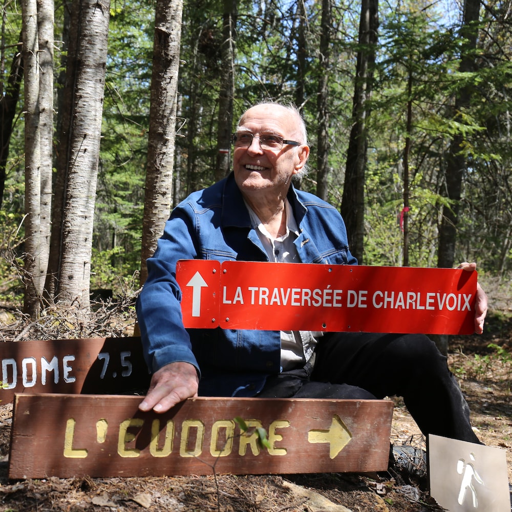 Un homme âgé dans une forêt, tient une affiche inscrit La Traversée de Charlevoix et une autre écrite L'Eudore. 