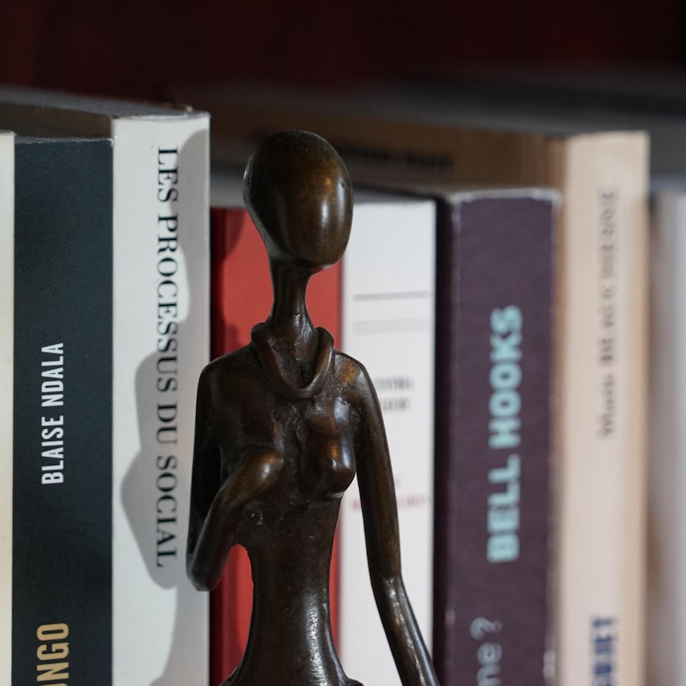 Une sculpture africaine en bois qui est un personnage féminin devant une étagère de livres.