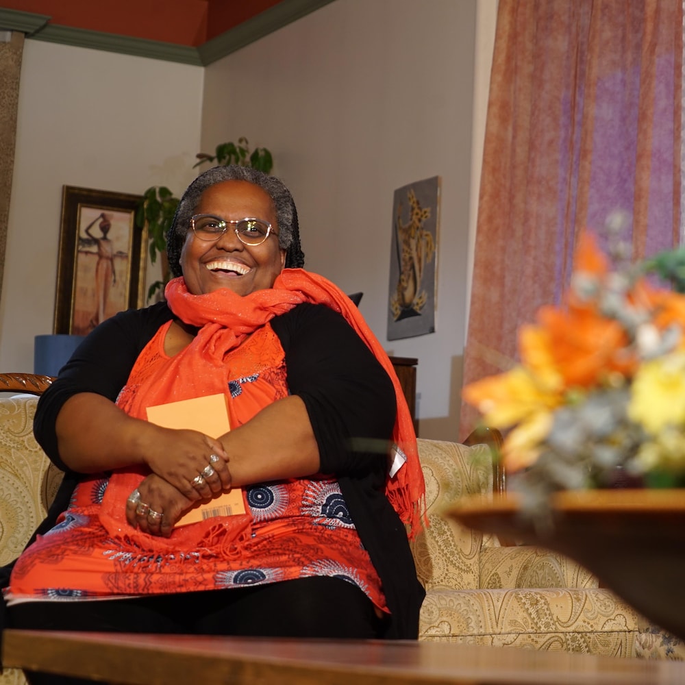 Jeanne-Marie Rugira est assise en souriant dans son salon avec un livre dans ses mains.
