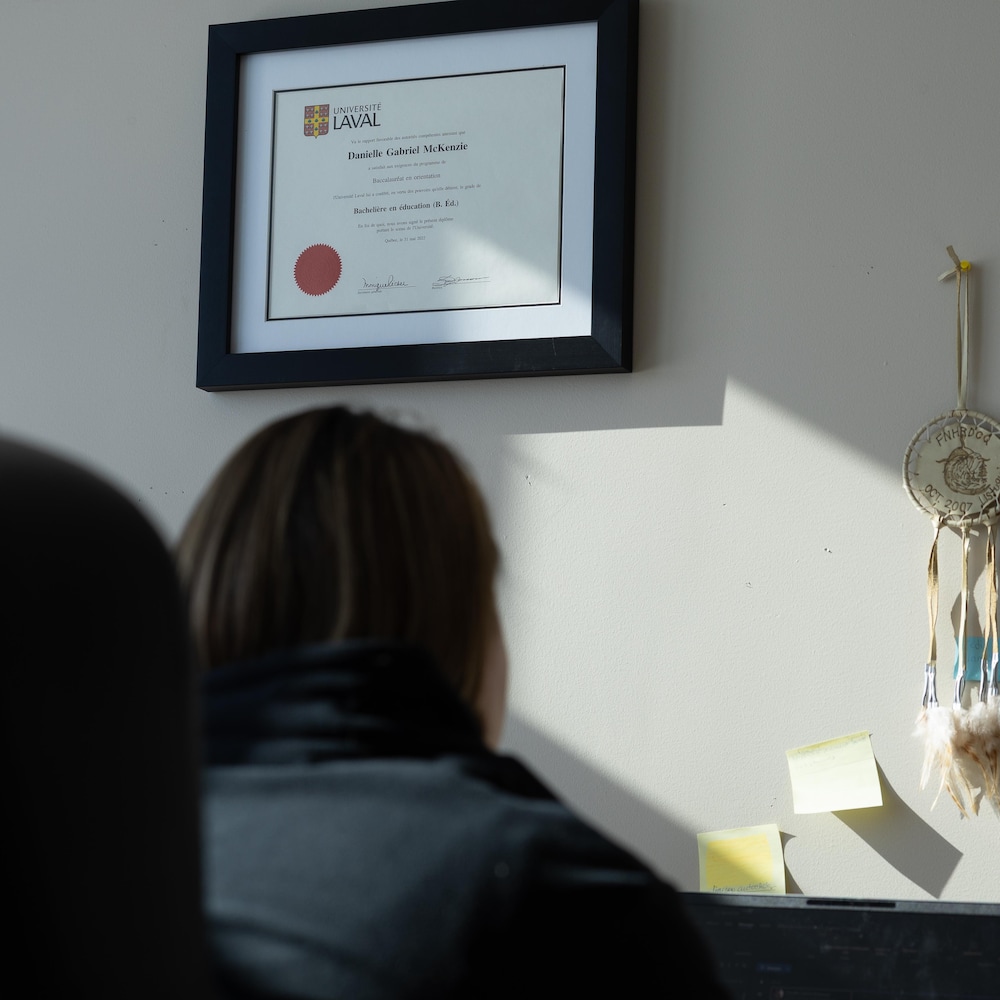 Une femme de dos assise à un bureau avec un diplôme universitaire au mur. 
