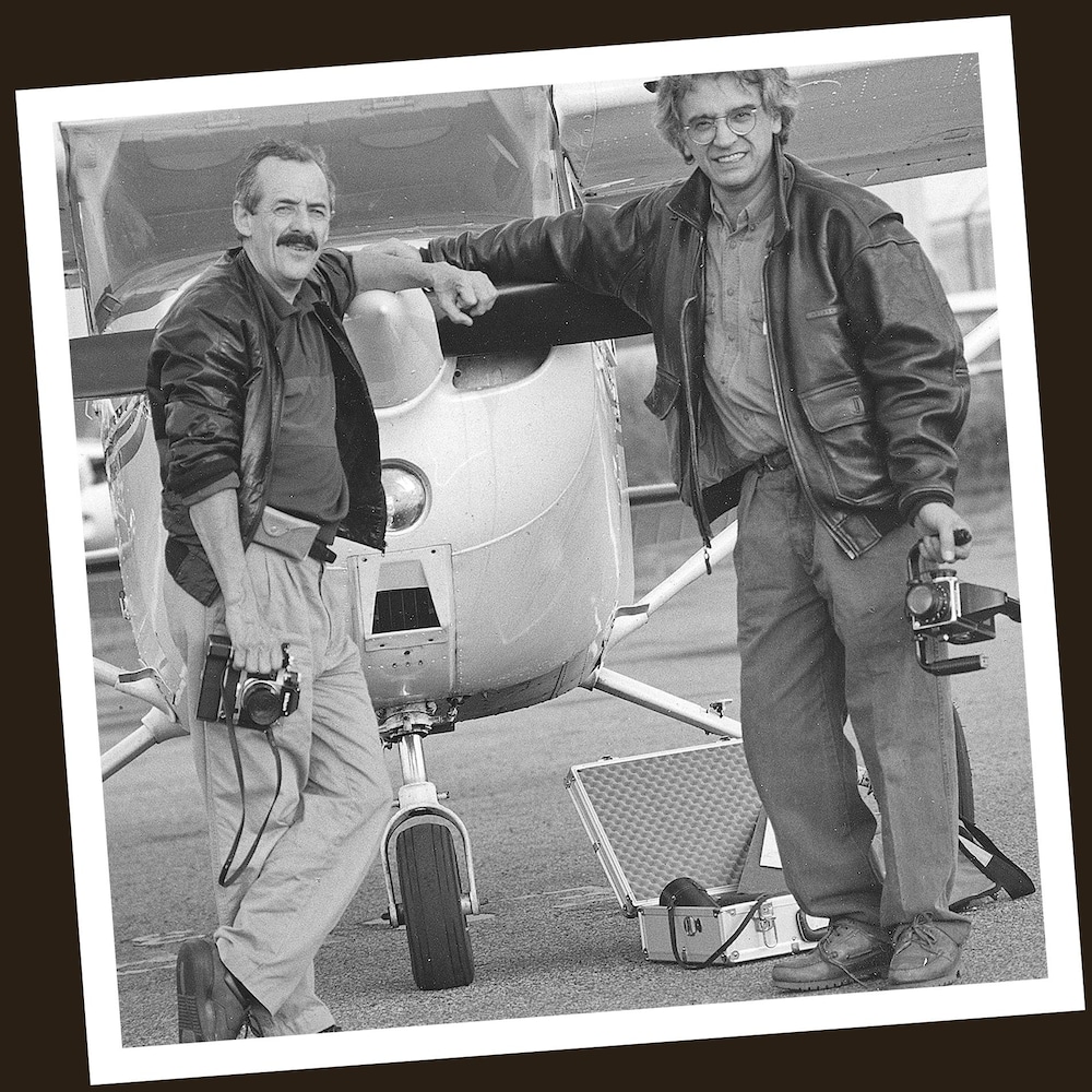 Deux hommes posent devant un petit avion avec des appareils photos dans leurs mains.
