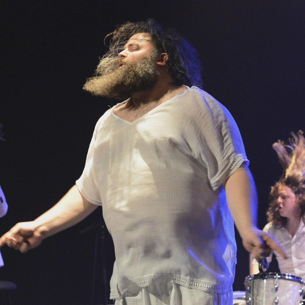 Un homme aux cheveux longs et à la barbe longue est vêtu de blanc. Il danse sur scène, les yeux fermés, devant un bassiste et un batteur qui se donnent autant.