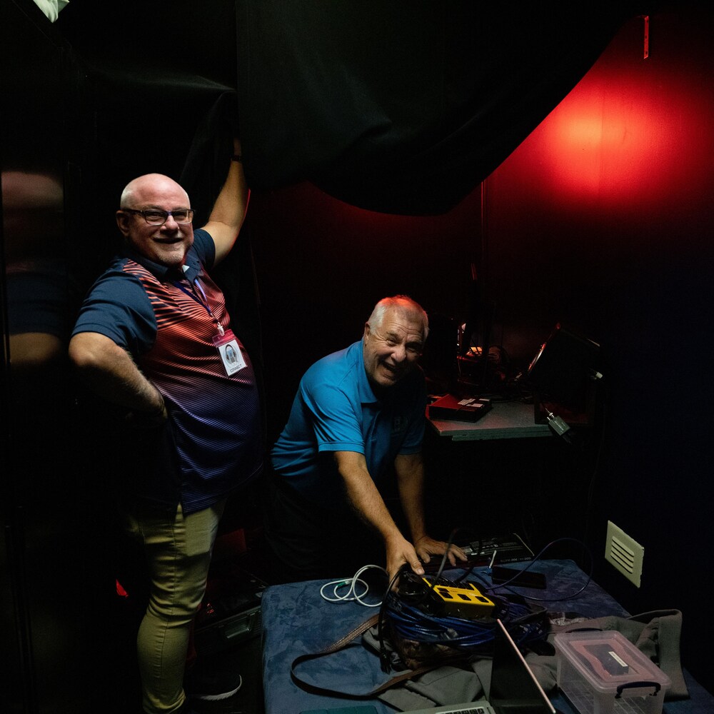 Deux hommes tout souriants  sont dans les coulisses d'un théâtre avec des files et des appareils électroniques. 