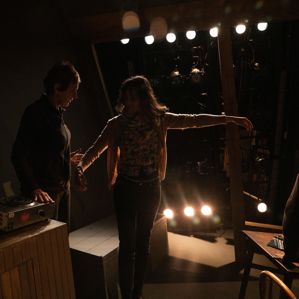 Une comédienne est avec un technicien en coulisse d'un théâtre sous un éclairage feutré. 