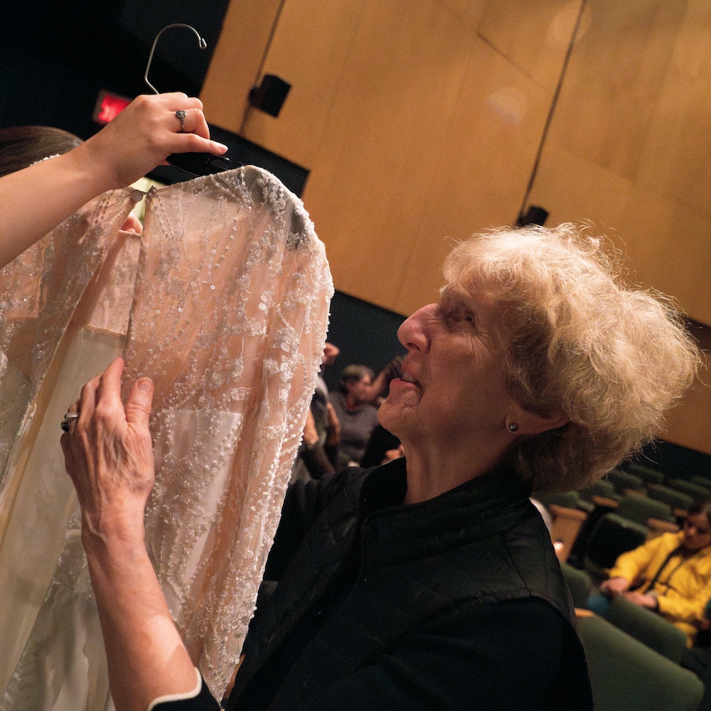 Une femme non voyante touche une robe de mariée sur un cintre qui est tenue par une femme dans une salle de spectacle.