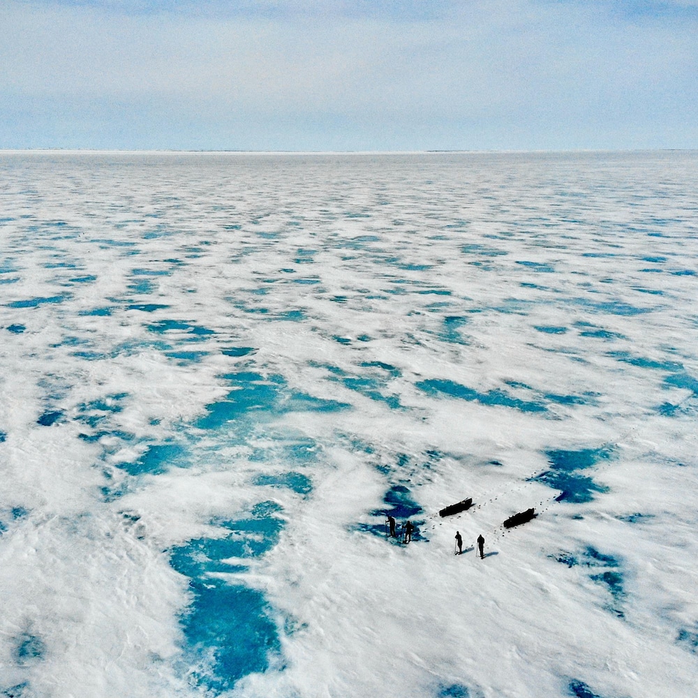 Vue d'un drone, en hauteur. On aperçoit quatre personnes qui tirent deux traîneaux. Le paysage est composé de glace, d'un bleu éclatant, et de neige à perte de vue.