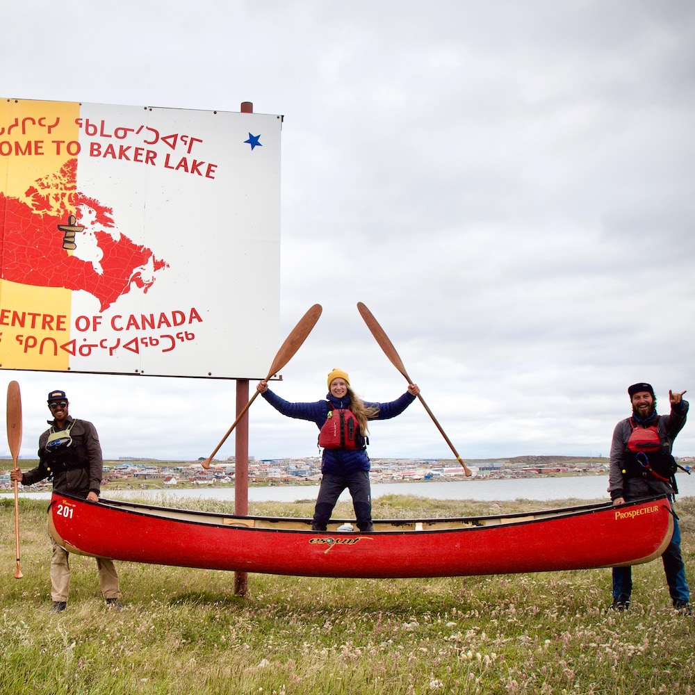 Les trois aventuriers et une femme posent avec leurs canots devant une affiche sur laquelle est inscrite « Bienvenue à Barker Lake, le centre du Canada »