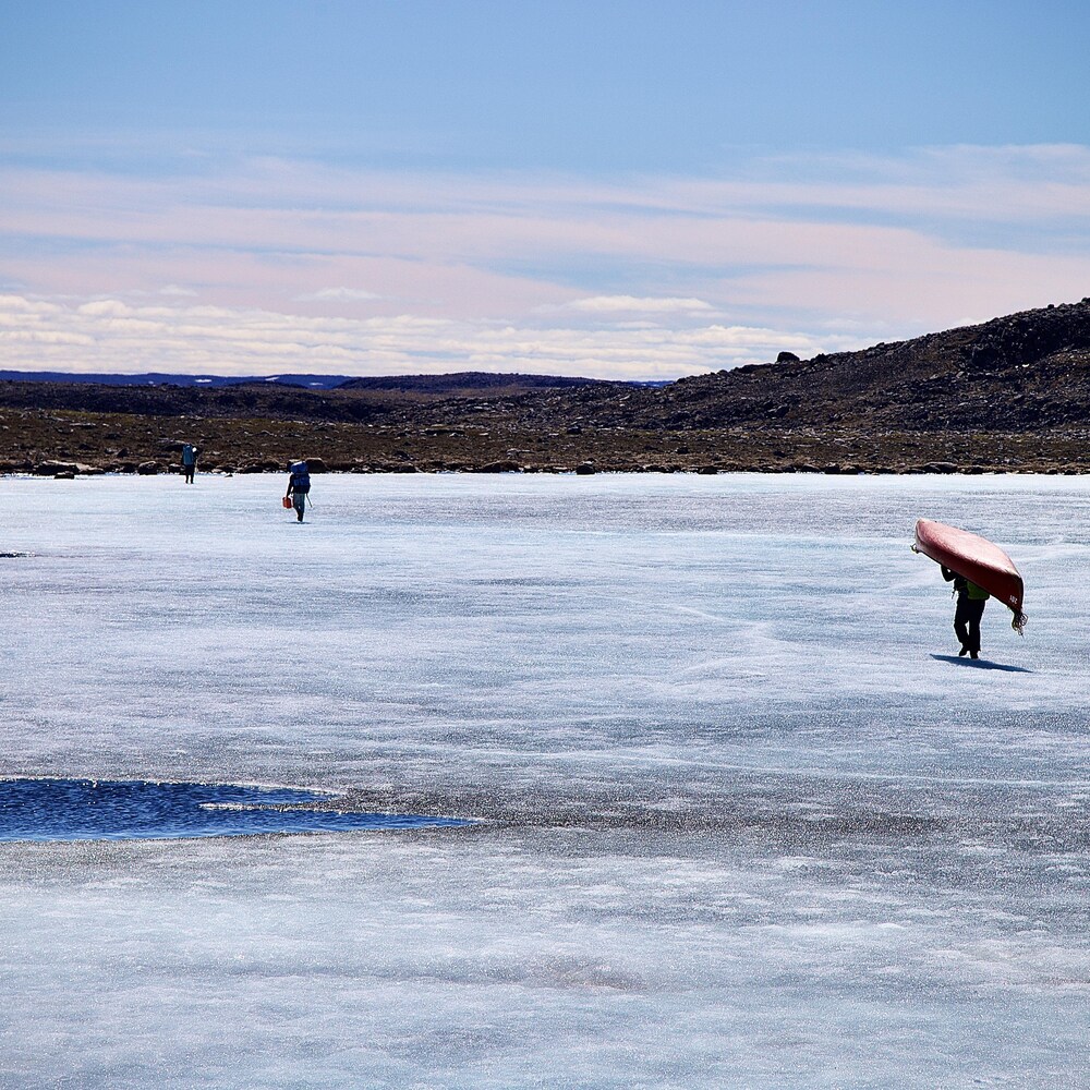 Trois hommes marchent sur un lac glacé, avec un canot et des vivres sur leur dos.
