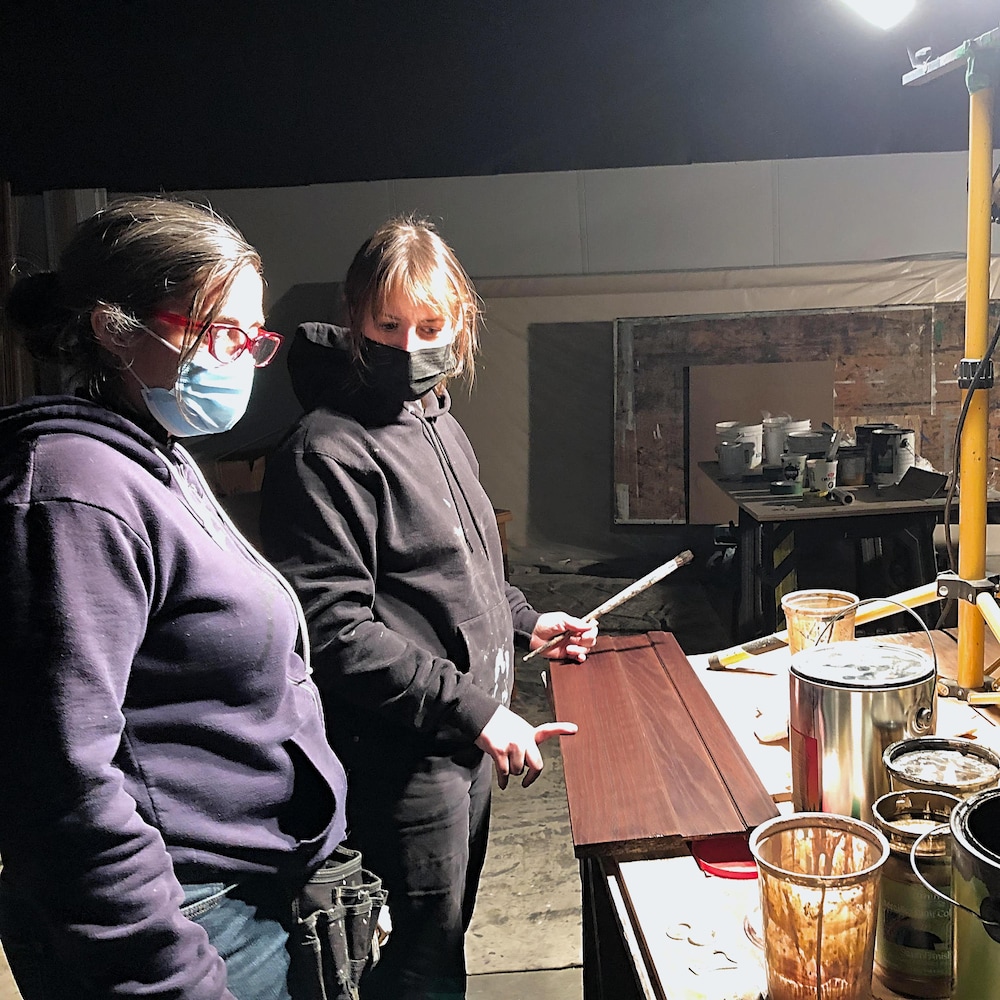 Deux femmes, à gauche de l’image. Elles sont devant une table sur laquelle il y a plusieurs gallons de teinture.