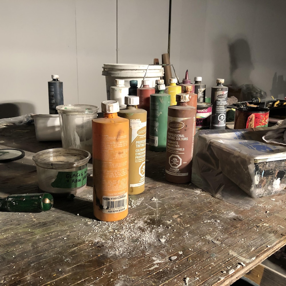 Sur une table en bois, des pots de peinture de différentes couleurs sont étalés dans le désordre. Du bouche-pores, une panne et quelques outils sont aussi sur la table.