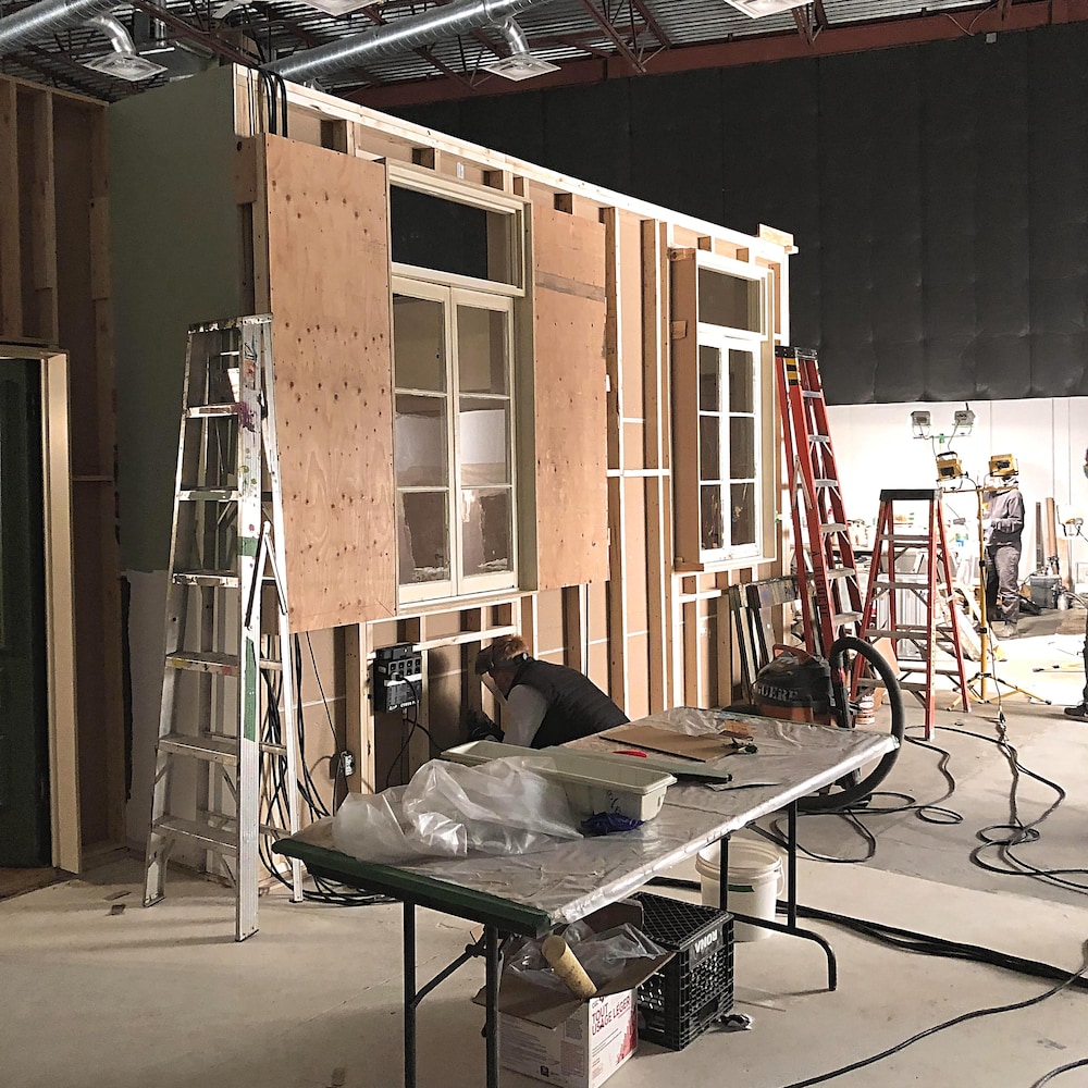 À l’arrière-plan, les faux murs en construction avec deux fenêtres antiques. Quelques escabeaux appuyés sur les faux murs. À l’avant-plan, une table sur laquelle des outils de travail sont déposés.