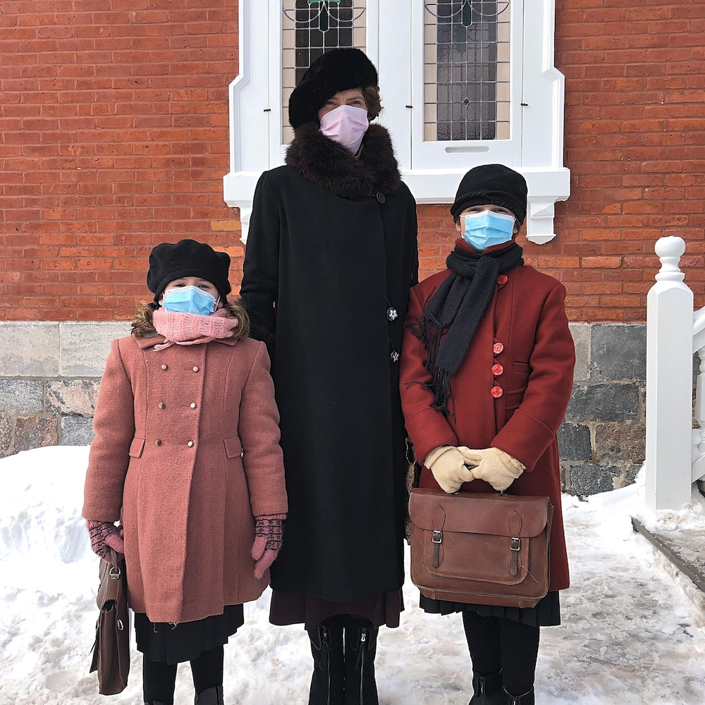 Hélène Florent, debout entre deux fillettes en âge de fréquenter une école primaire, est devant la maison de la rue des Ursulines. Les fillettes ont chacune un vieux sac de cuir à la main. Les trois portent un masque médical.