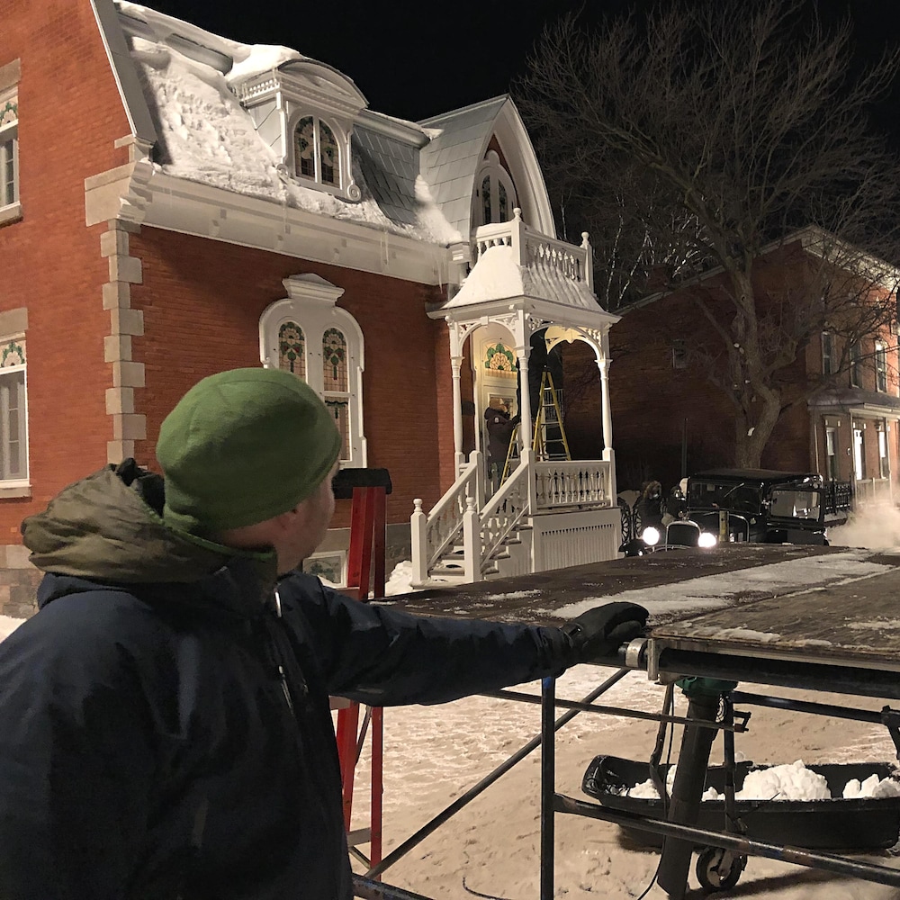 Image de soir. À l’avant, à gauche, un homme de profil pose sa main gauche sur un échafaud servant à l’équipe de production. Il regarde vers la maison où l’équipe technique ajuste une lampe sur le balcon en prévision d’une scène qui sera tournée quelques minutes plus tard.