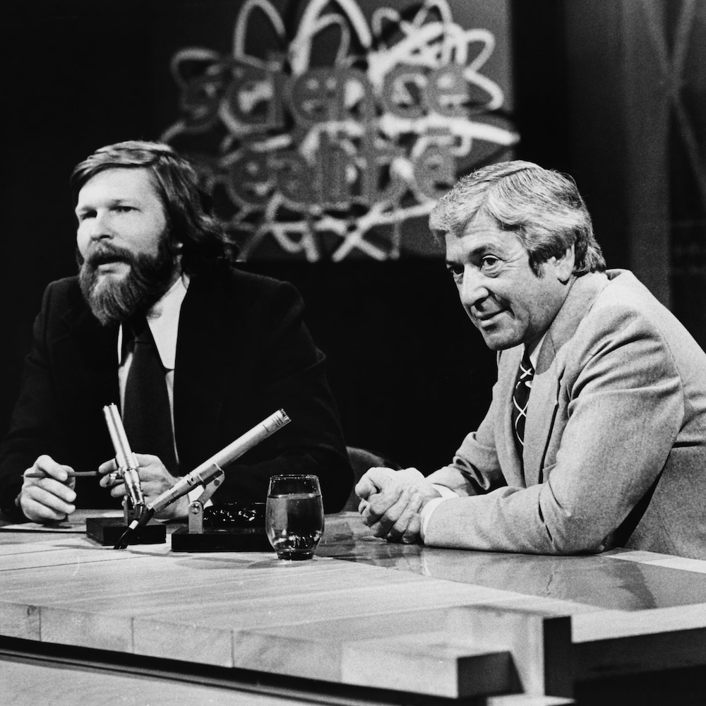 Dans un studio de télévision, Joël Le Bigot et Fernand Seguin assis à une table surmontée de deux microphones. En arrière-plan, on voit le titre de l'émission dans un écran sur le mur