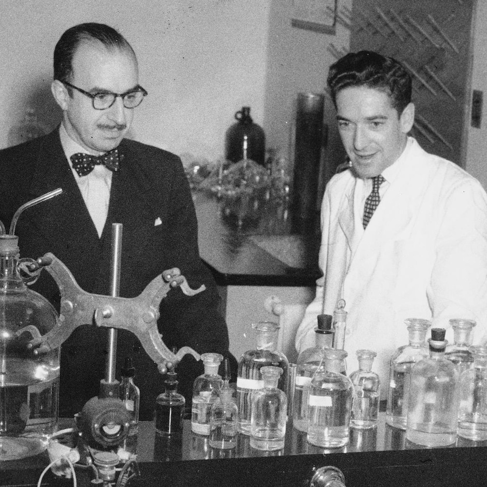 Dans un laboratoire, l'animateur Fernand Seguin derrière une table remplie de fioles. À ses côtés, le Dr Paul Dumas.