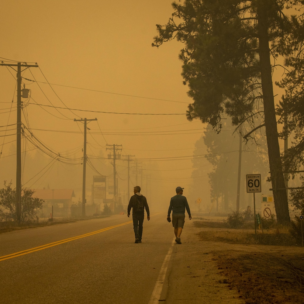 Deux hommes marchent le long d’une route alors que la fumée des feux est très épaisse, à Scotch Creek, en Colombie-Britannique, le samedi 19 août 2023.