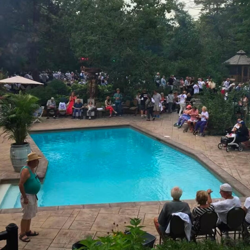 Des gens sont rassemblés dans une cour arrière près d'une piscine. Une grande foule est présente, certains mangent, d'autres boivent de la bière.