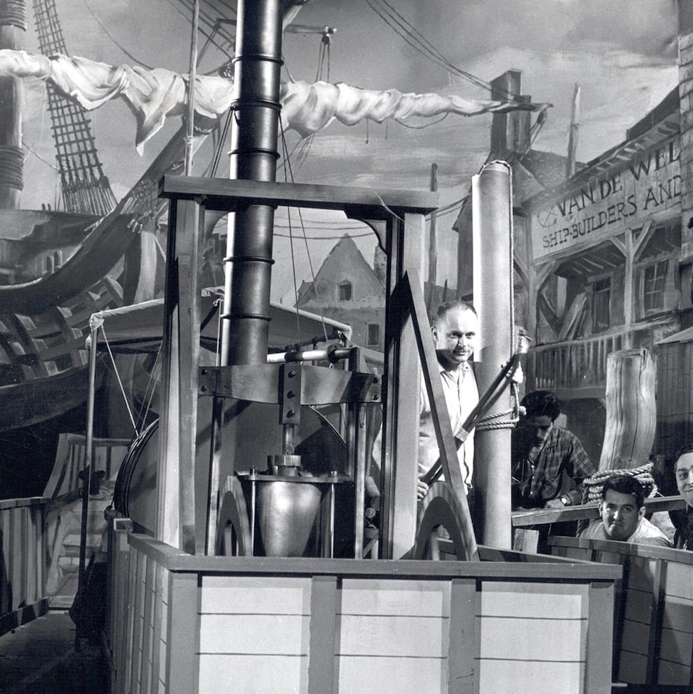Frédéric Back à bord d'un décor de bateau avec trois collègues à ses côtés. À l'arrière-plan, une grande toile qui montre l'illustration d'un port complète le décor.