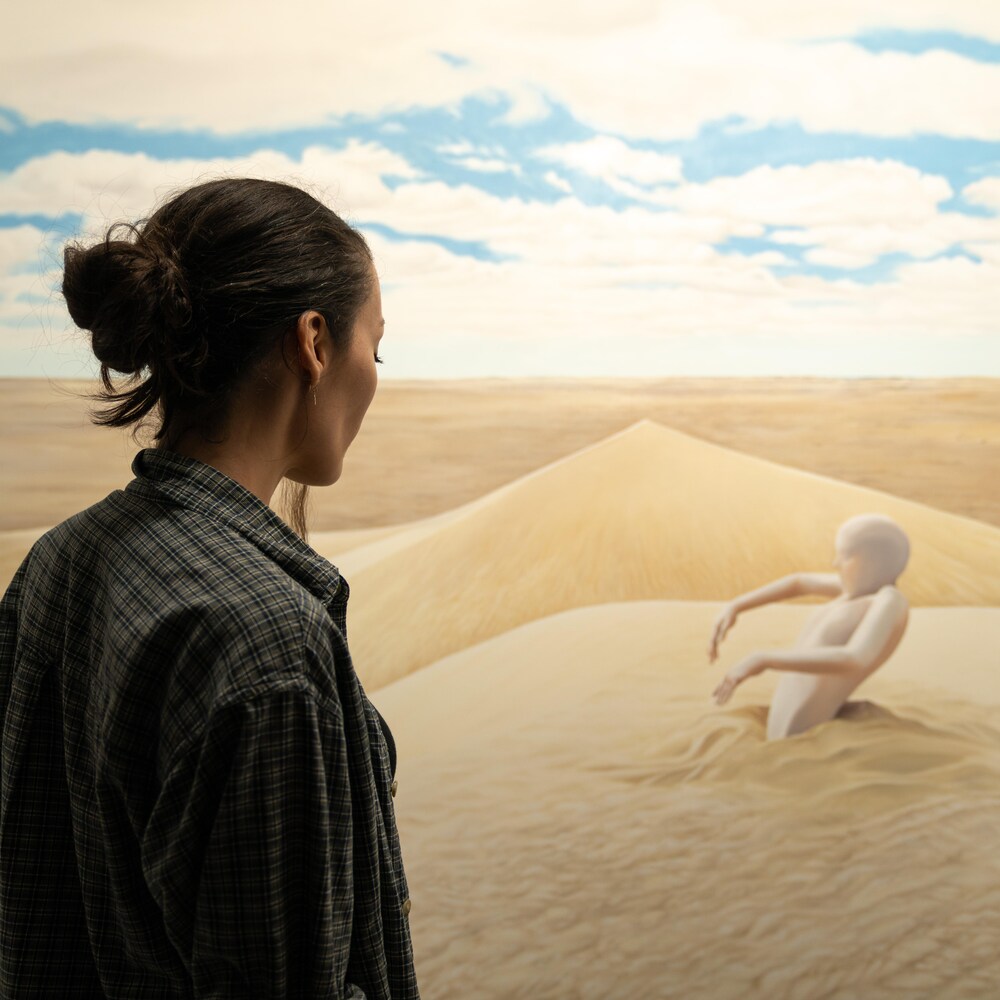 Mireille, de dos, regarde sa toile. On y voit des dunes et une forme humaine beige qui sort du sable.