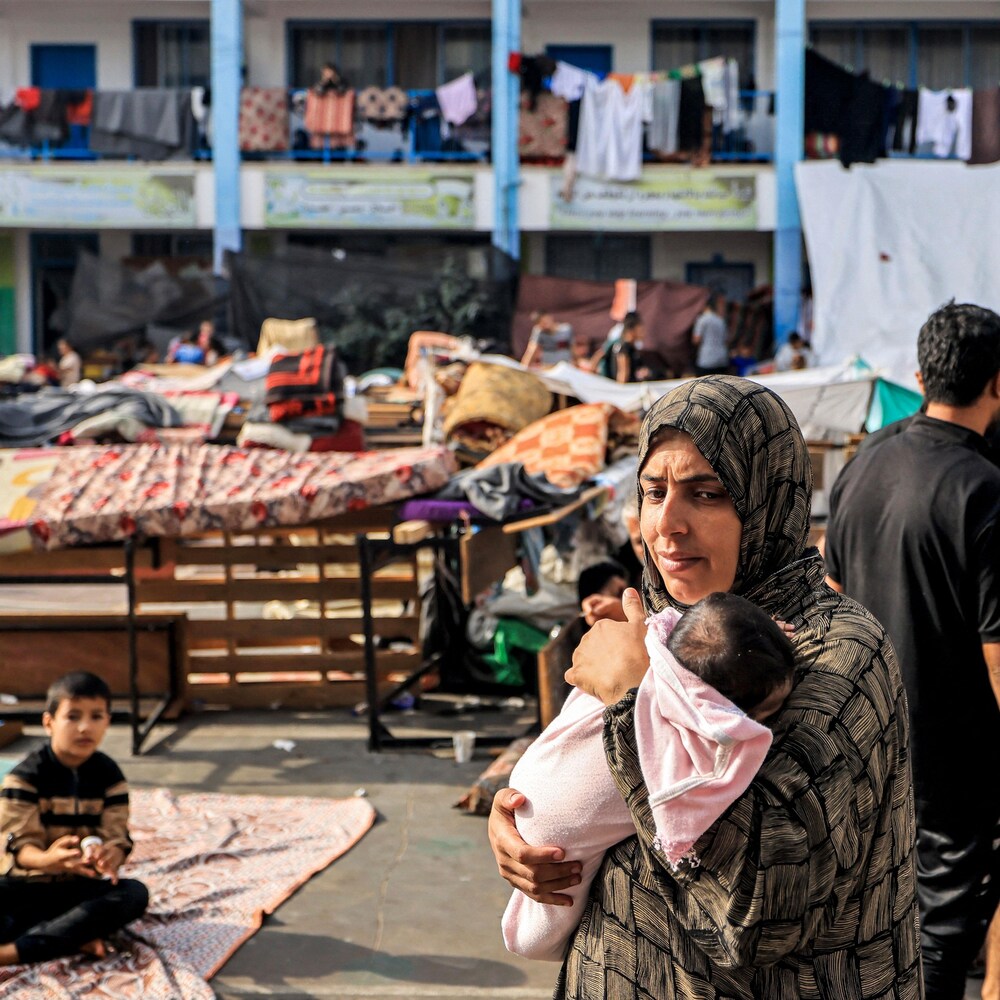 Une mère tenant un bébé marche au milieu de lits et de personnes dans le désordre d'un camp de réfugiés.