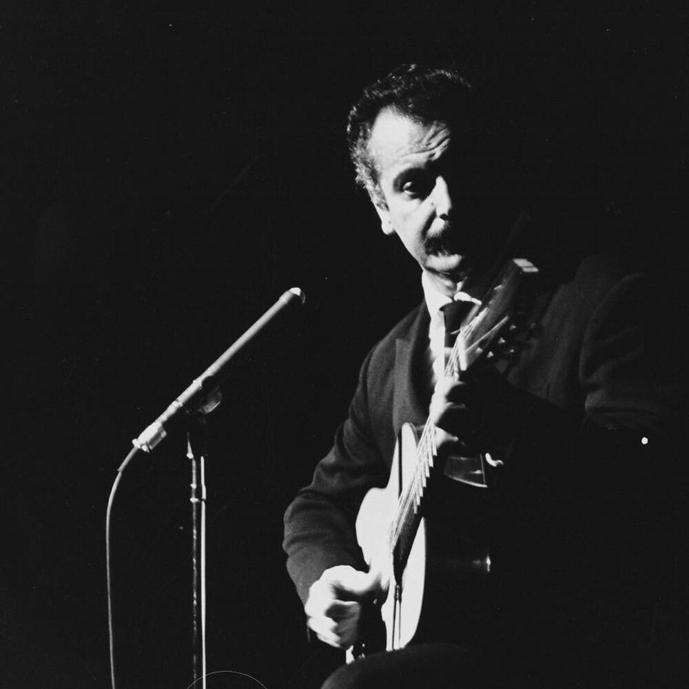 Le chanteur français Georges Brassens debout sur la scène en s'accompagnant à la guitare.