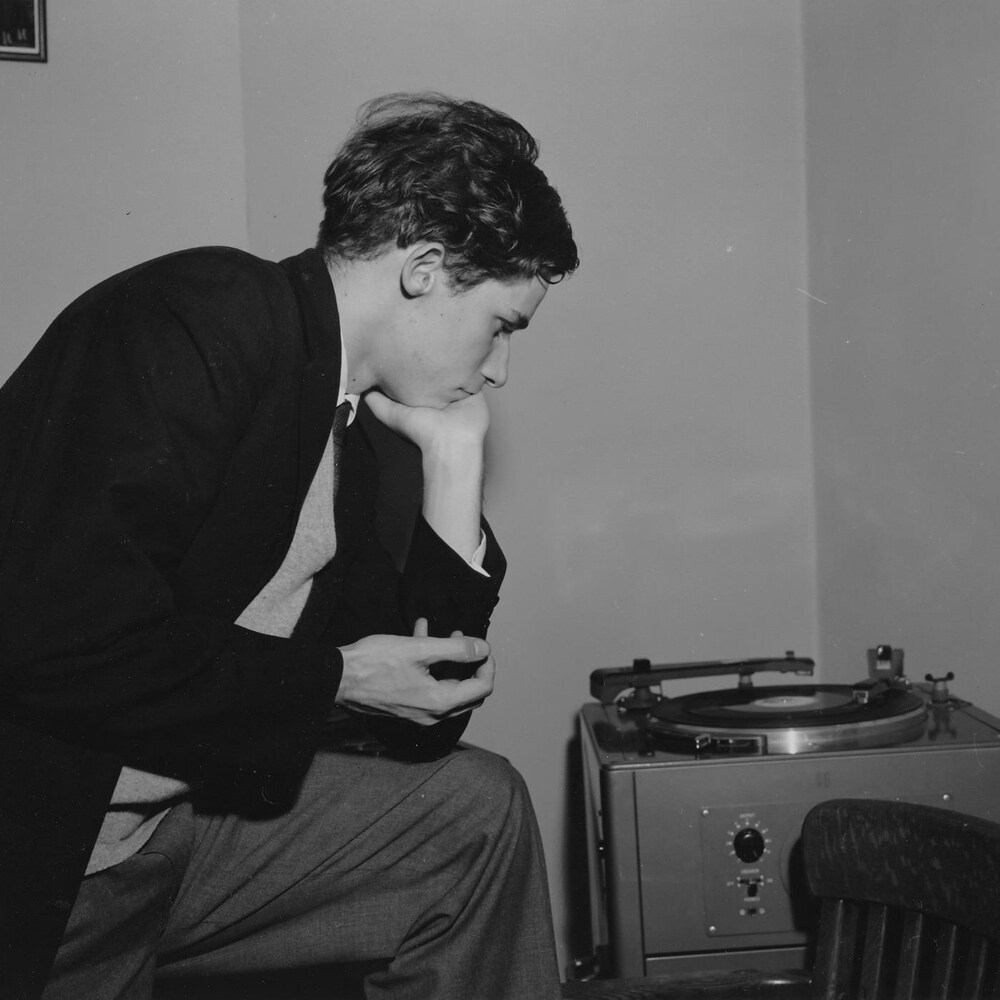 Glenn Gould dans un studio, accroupi près d'un tourne-disque qui semble écouter attentivement.