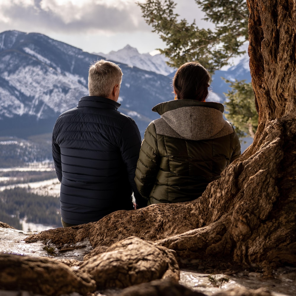 Un homme et une femme sont assis de dos sur les énormes racines d'un arbre et regardent les sommets des montagnes.