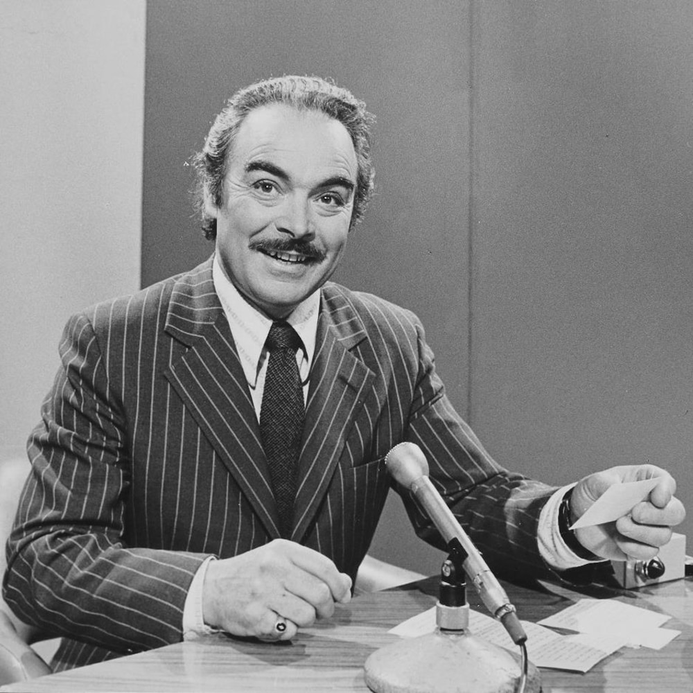Henri Bergeron, carton à la main, animant assis à un bureau devant un micro sur pied.
