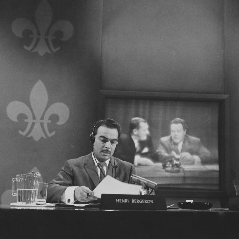 Henri Bergeron, assis à un bureau devant un décor fleurdelisé.