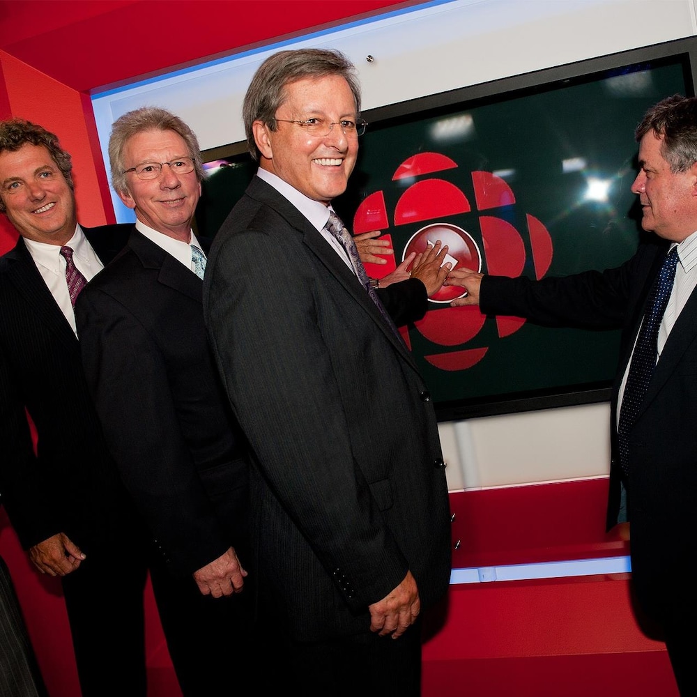 Sylvain Lafrance, Jean-Pierre Blackburn, Jean Tremblay et Michel Gagné touchent un écran avec le logo de Radio-Canada en souriant.