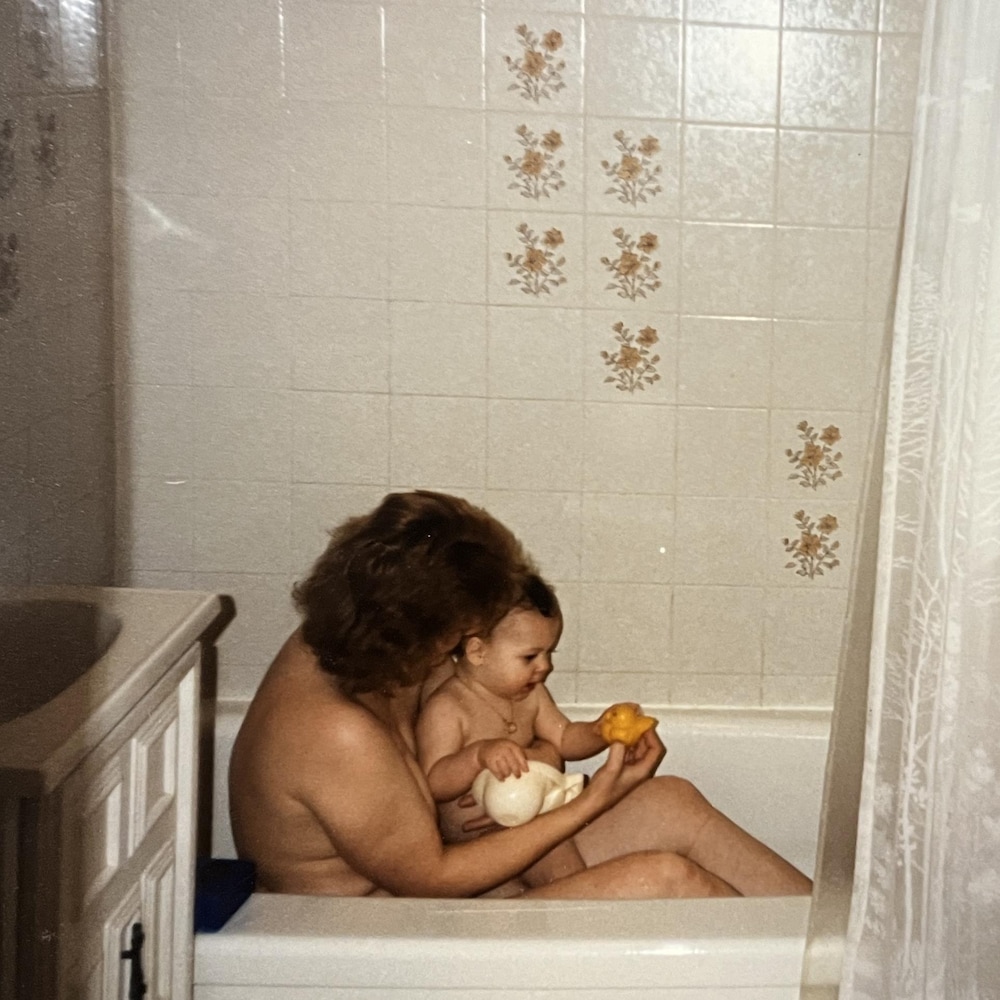 Francine Turbide tient Isabelle Rivest dans ses bras dans le bain. Bébé, Isabelle Rivest peine à se tenir debout et tend la main vers un canard en plastique.