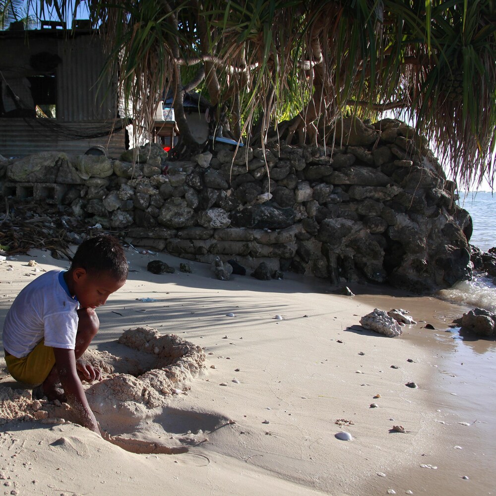 L'enfant joue dans le sable près de sa maison, qui est construite à côté de l'eau et entourée d'un muret de pierre.