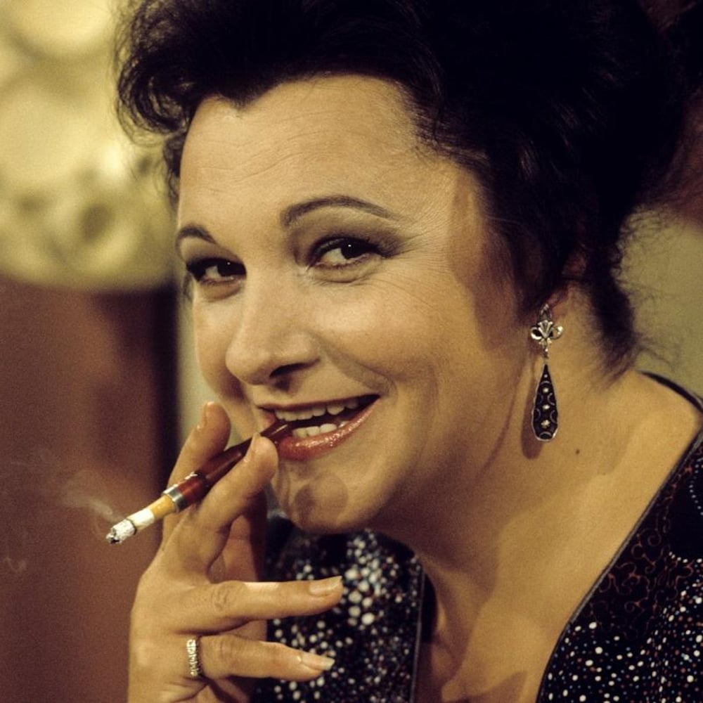 Dans un studio de télévision, l'animatrice Lise Payette est assise à son pupitre et tient un fume-cigarette contenant une cigarette allumée entre ses lèvres.