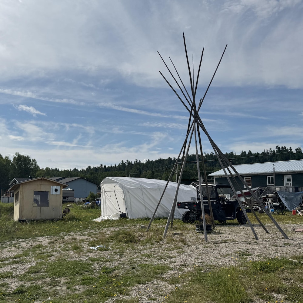 Une structure formée de longues perches de bois se dresse près de maisons, d’une tente blanche et d’un véhicule tout-terrain.