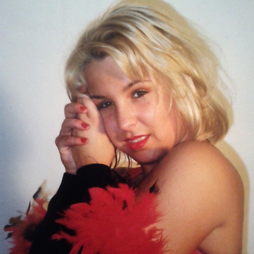 Une jeune femme blonde porte des vêtements rouge et noir et elle porte autour de ses bras des plumes des mêmes couleurs.