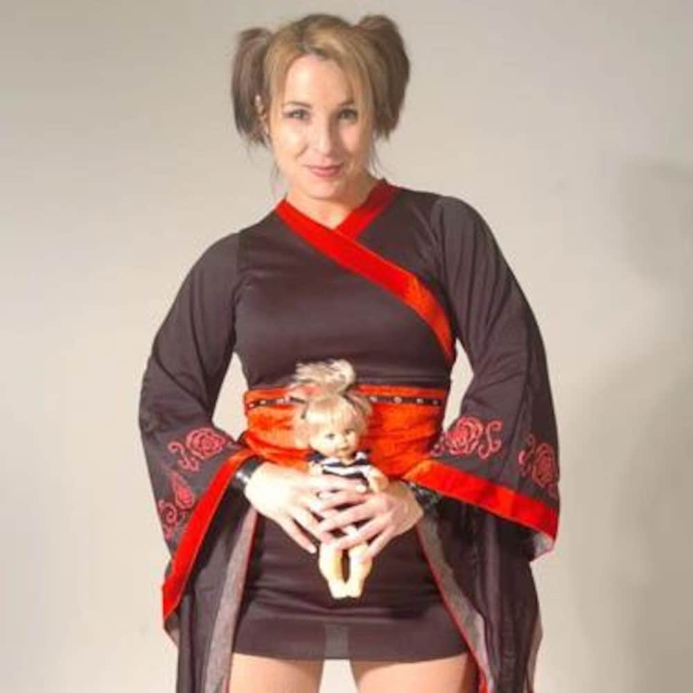 Une femme porte un vêtement qui ressemble à un kimono et tient une poupée blonde dans ses mains.