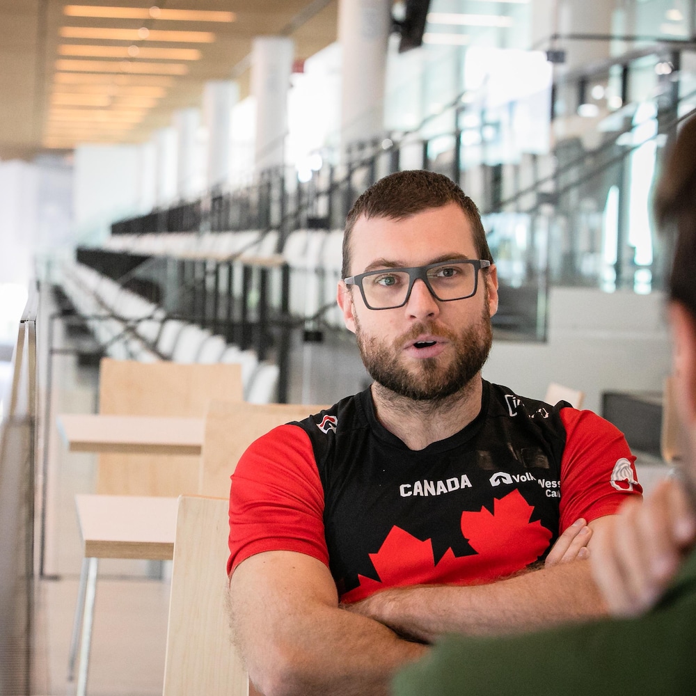 Laurent Dubreuil, portant des lunettes et un chandail d'Équipe Canada, est assis à une table devant une patinoire intérieure.