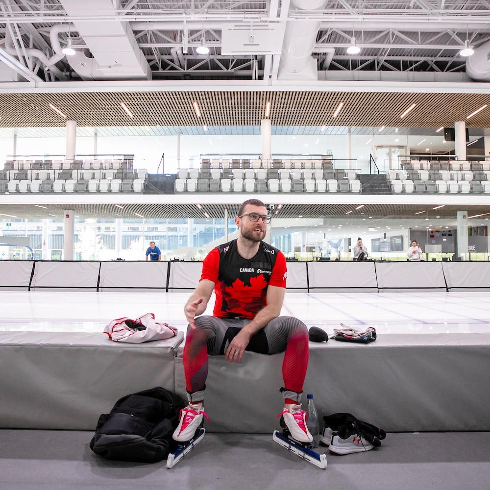 Laurent Dubreuil est assis sur un banc de protection au bord de la glace, ses patins de vitesse aux pieds, et discute avec une personne hors champ dans un centre de patinage moderne.