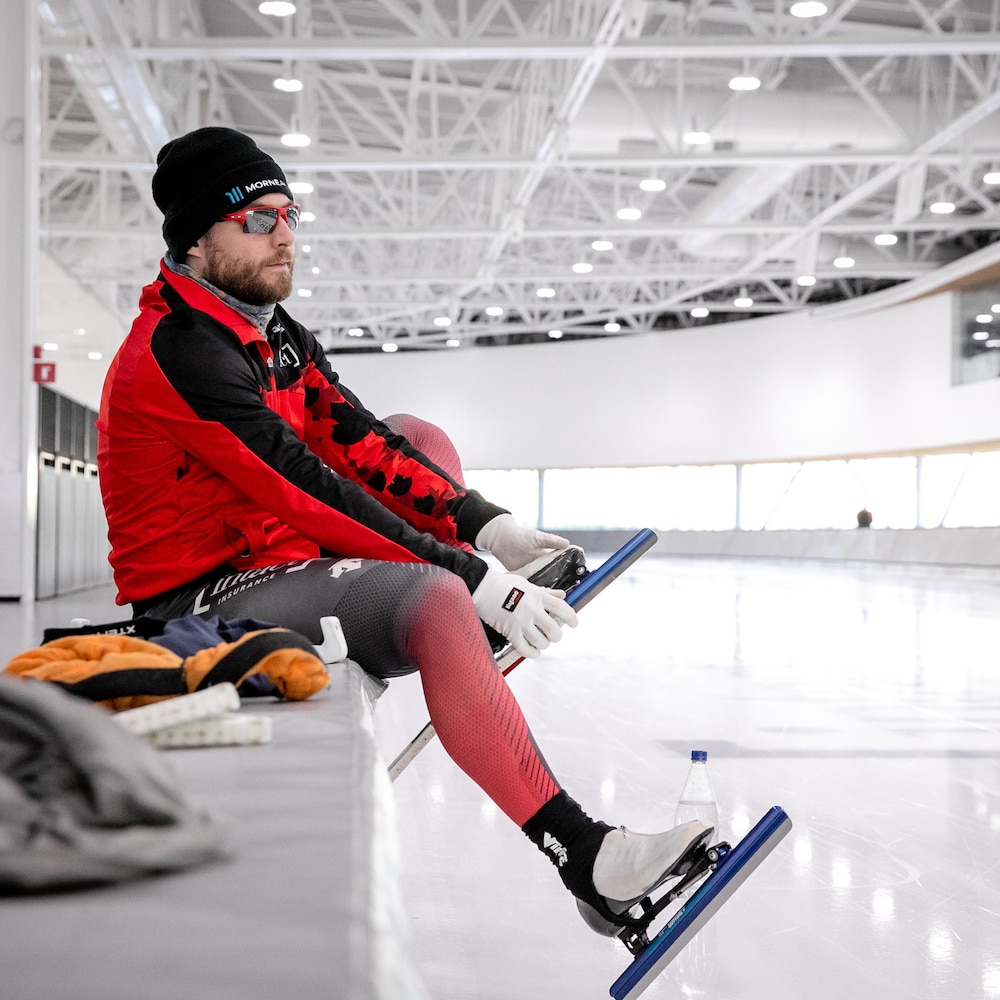 Laurent Dubreuil, assis au bord d'une patinoire intérieure, ajuste ses patins de vitesse tout en observant la glace.