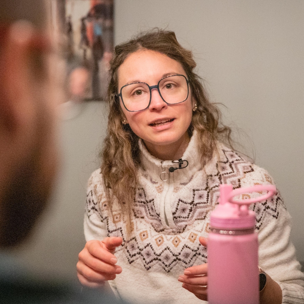 Une femme portant des lunettes et un chandail à motifs parle activement, assise à une table avec une bouteille rose devant elle.