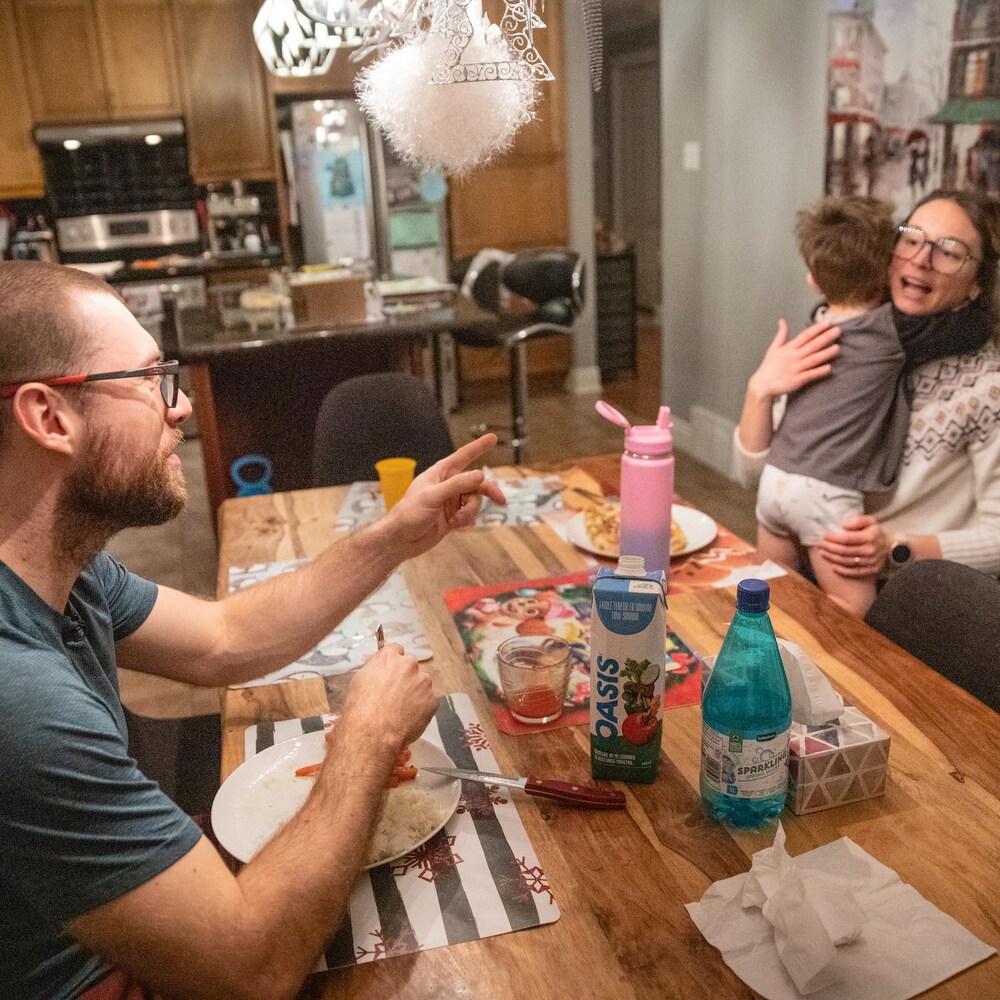 À table, Laurent Dubreuil pointe son doigt vers sa conjointe qui tient leur jeune fils dans ses bras.