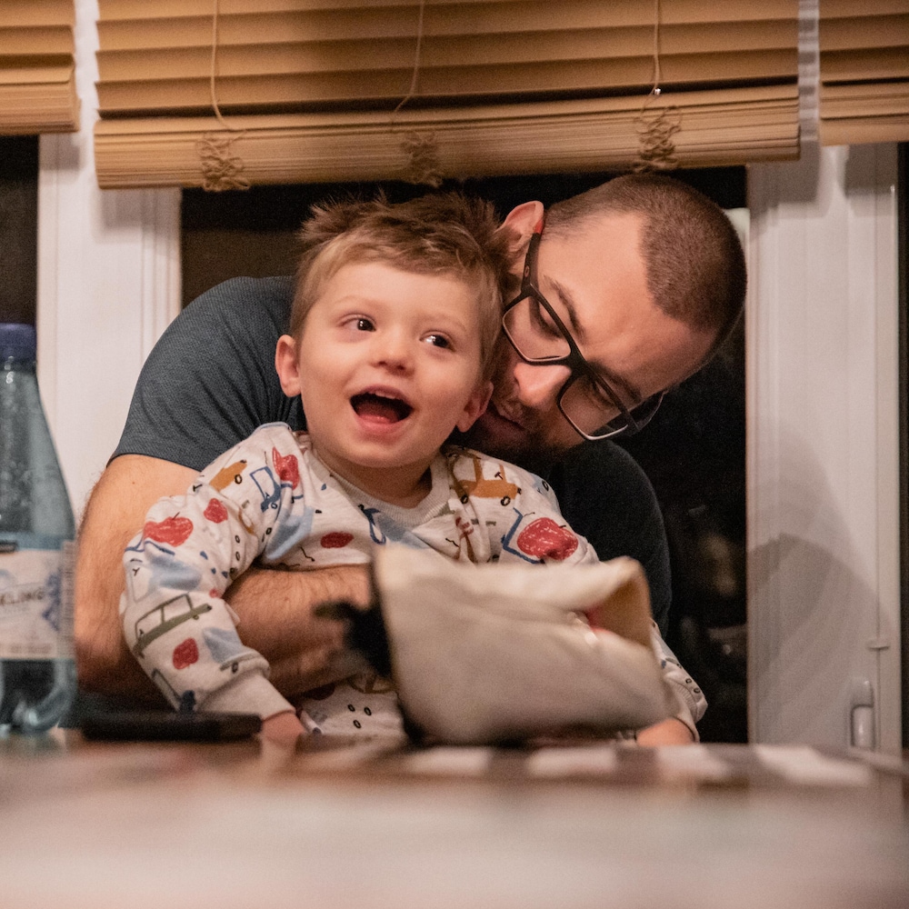Laurent Dubreuil tient son jeune fils souriant dans ses bras, assis à une table devant une fenêtre aux stores baissés.