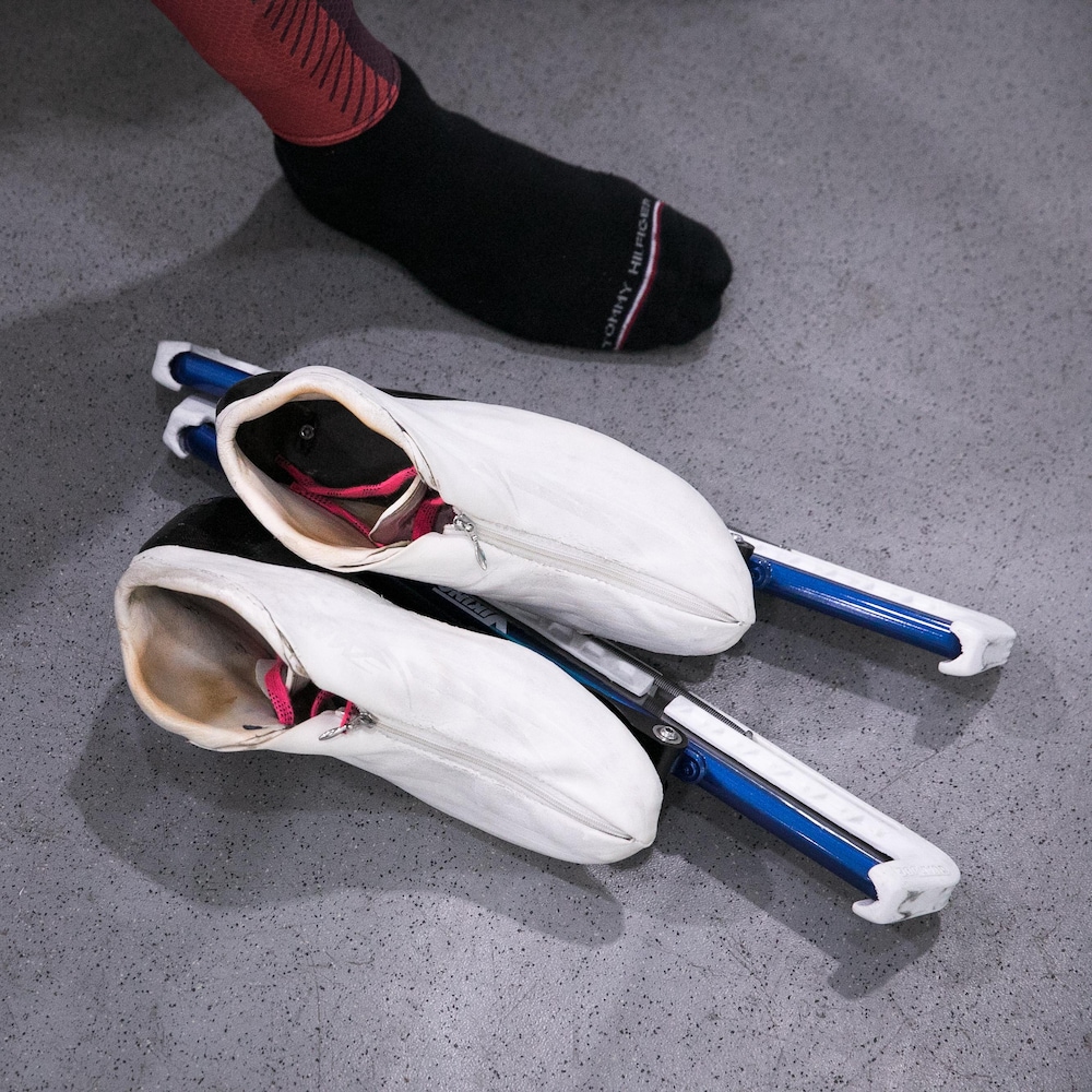 Une paire de patins de vitesse blancs posée sur le sol à côté d'un pied vêtu d'une chaussette noire.