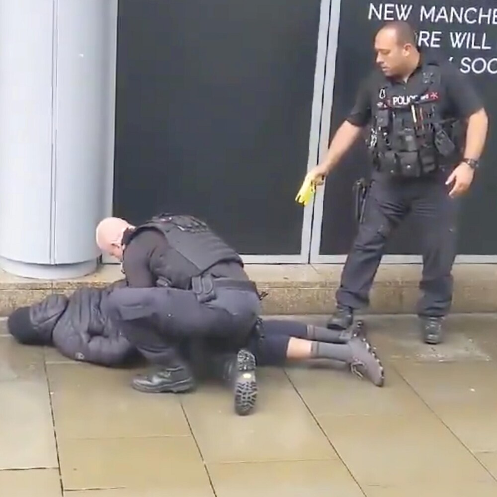 Un policier retient un homme contre le sol, un deuxième policier pointe un pistolet paralysant dans sa direction.