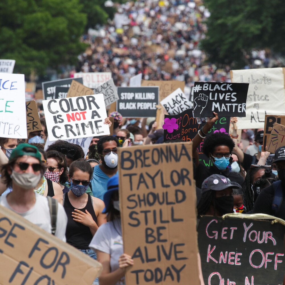 Une marée bigarrée de manifestants portant le masque et des pancartes occupent toute l'image.