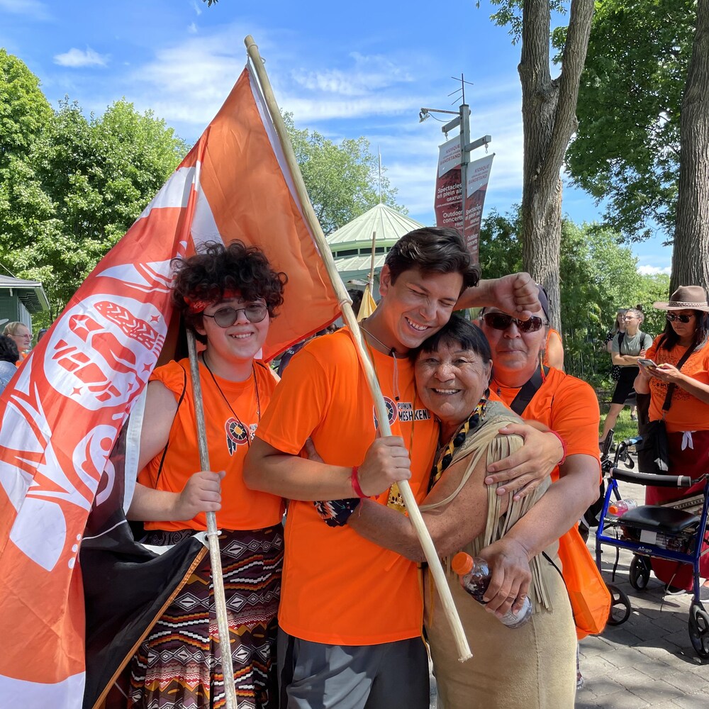 Des personnes vêtues de gilets oranges tiennent une banderole où il est écrit « Every Child Matters ». Au centre, Jay Launière-Mathias, serre sa grand mère dans ses bras.