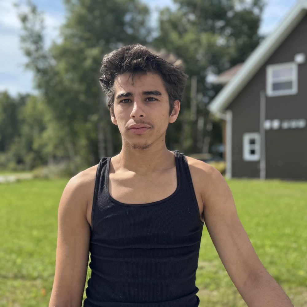 n jeune homme vêtu d’un débardeur noir se tient sur une pelouse, devant un bâtiment bordé par la forêt.