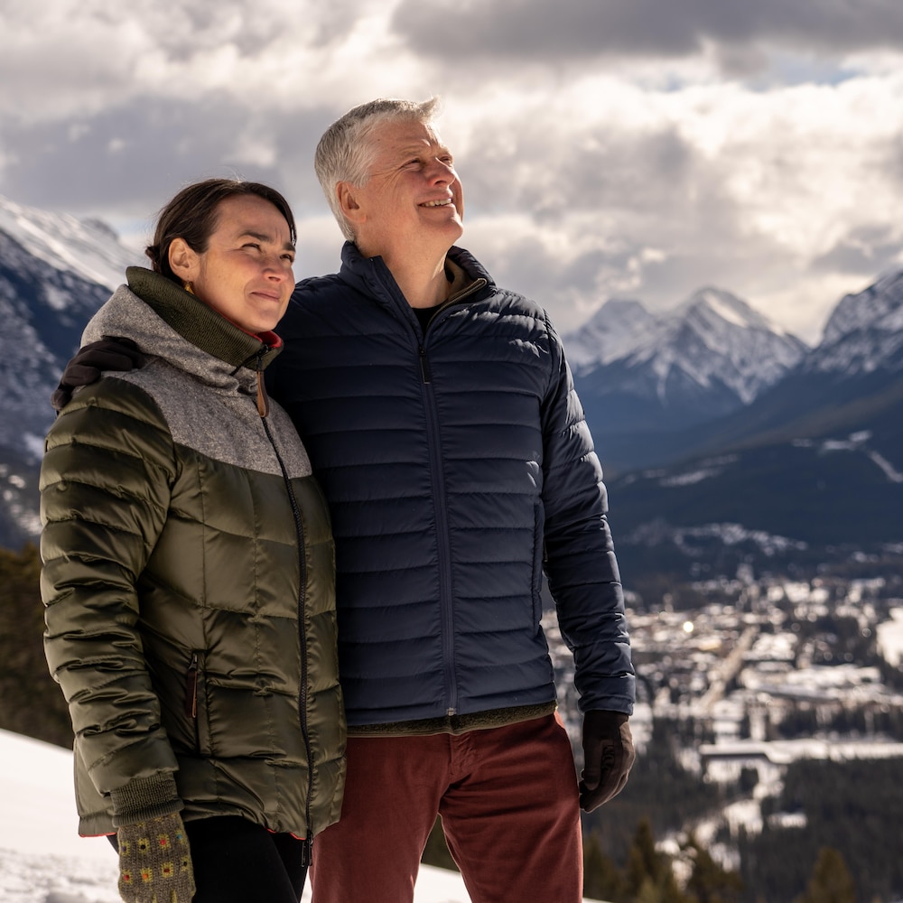 Une femme dans la quarantaine et un homme dans la cinquantaine en habits d'hiver posent devant un décor magnifique de sommets de montagne. Ils sont souriants et leur regard est vers le ciel. 